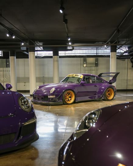 Purple RWB Porsche 911 with gold wheels, body kit, and large spoiler, flanked by other purple cars.
