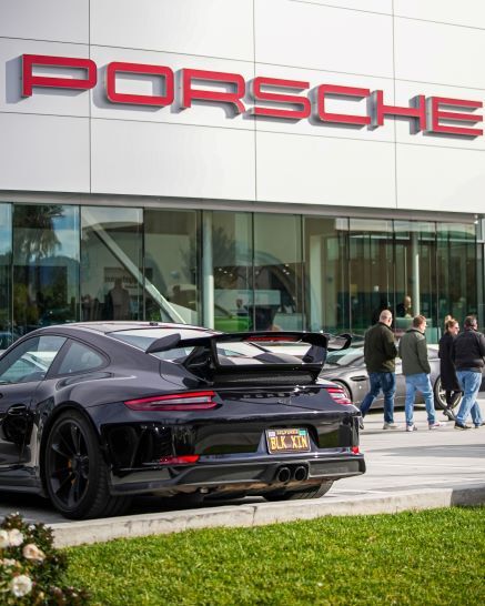 A black Porsche with a large spoiler and personalized license plate BLK XTN parked in front of a Porsche dealership.