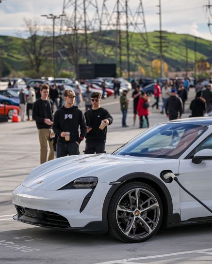 A silver Porsche Taycan electric car charging in a busy parking lot with people and power lines in the background.