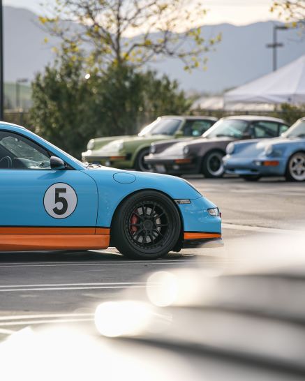 A light blue Porsche with an orange stripe and the number 5 on its side, parked among other Porsches.