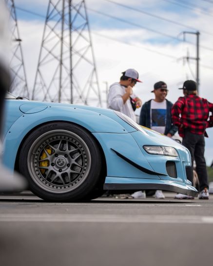 A light blue custom sports car with large silver rims and yellow brake calipers, with three people in the background.