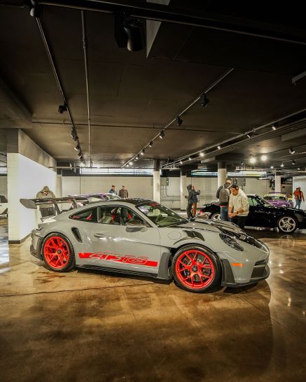 A grey Porsche 911 GT3 RS with red wheels and decals on a polished concrete floor in a showroom with people.