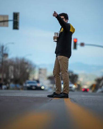 Young man in a black and yellow hoodie and tan pants holding a coffee cup, pointing across a street.