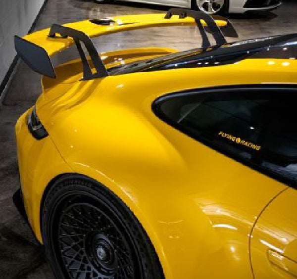 A yellow sports car with a large rear spoiler is parked in a garage, with a white sports car in the background.