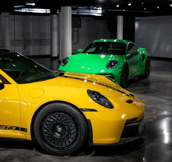 Front of a yellow Porsche 911 GT3 with a black wheel, and a green Porsche 911 in the background.