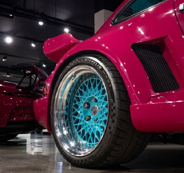 Close-up of a hot pink sports car's rear wheel with a vibrant turquoise, intricate mesh design.