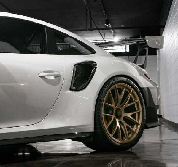 Rear of a white Porsche sports car with gold wheels, large spoiler, and air vents, in a garage.