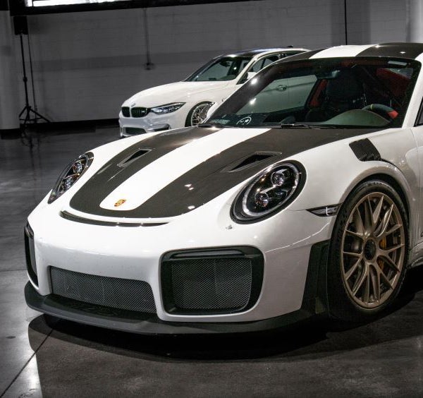 Front of a white Porsche race car with black stripes on hood and roof, and a white BMW in the background.