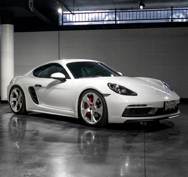 A white Porsche Cayman GT4 with red brake calipers is parked on a shiny dark floor in a garage.