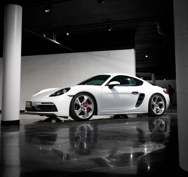White Porsche Cayman sports car on a reflective concrete floor, with white walls and dark ceiling.