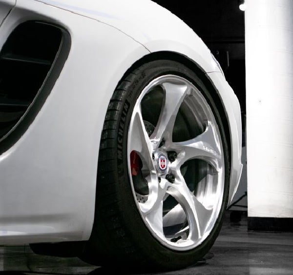 Close-up of a white sports car’s front fender and wheel, highlighting the chrome rim with red brake calipers.