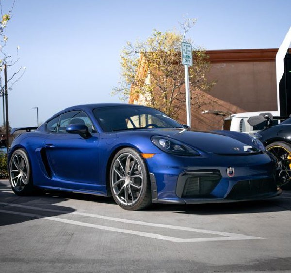 A blue Porsche 718 Cayman GT4 parked in a parking lot, viewed from the front quarter angle on a sunny day.