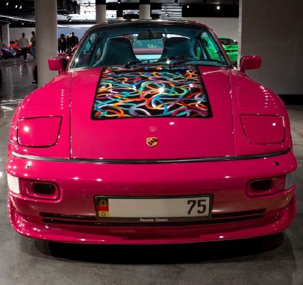 Front view of a shiny, hot pink Porsche with a black rectangular hood insert featuring colorful wavy lines.