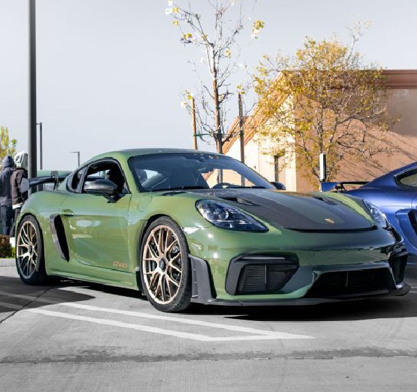 A military green Porsche 718 Cayman GT4 RS with bronze wheels and black hood parked in a lot.