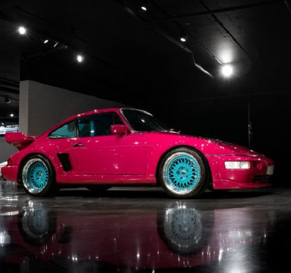 A bright pink Porsche with vivid turquoise wheels, parked on a reflective black floor in a dimly lit room.