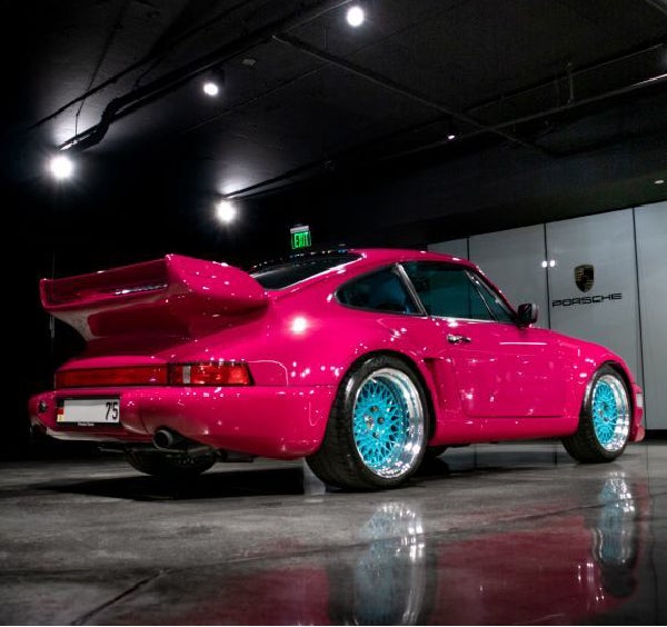 Rear view of a bright pink Porsche 911 with a large rear spoiler and turquoise blue wheels.