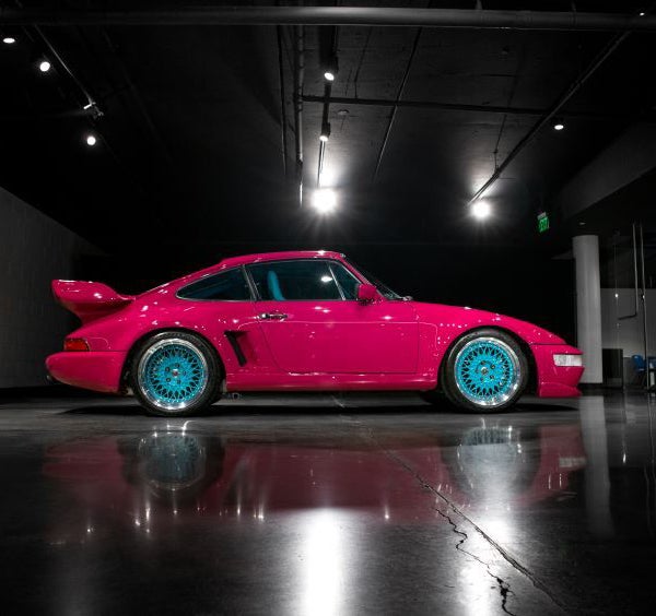 Side view of a bright pink Porsche with turquoise wheels and a large rear spoiler, parked on a reflective black floor.