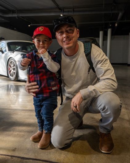 A father and young son smile for a photo in front of a white Porsche in an indoor parking garage.