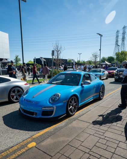 A bright blue Porsche with racing stripes is parked at a car show surrounded by people and other vehicles.