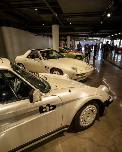 Vintage Porsche cars are displayed in a brightly lit showroom with people viewing them.