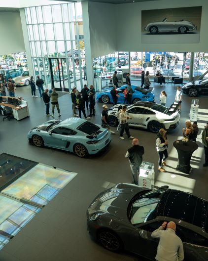 People browse a showroom filled with various Porsche cars displayed on a polished floor.