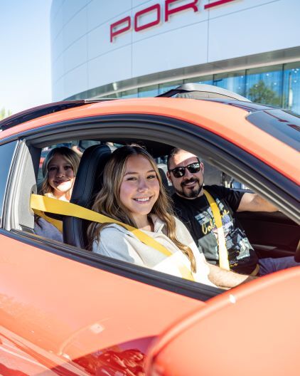 A family of three smiles while seated inside a red Porsche, with the Porsche logo visible in the background.