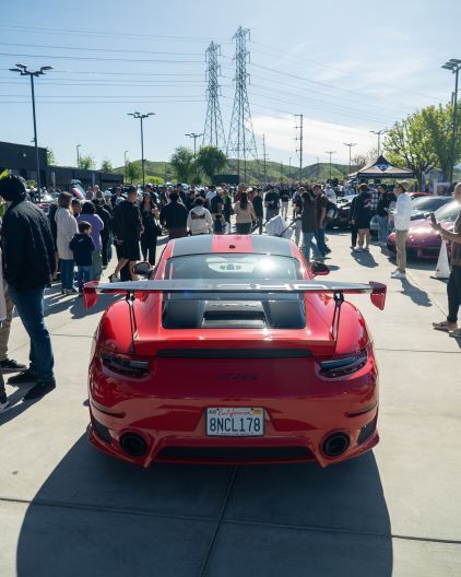 A red Porsche with a large rear wing sits parked at a car show surrounded by a crowd of onlookers.
