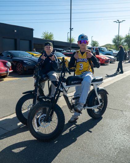 Two people wearing sunglasses pose on electric motorcycles in front of parked luxury cars.
