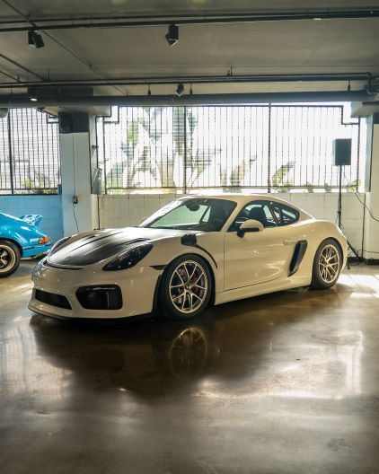 A white Porsche Cayman coupe sits in a parking garage with a blue car visible to the left.