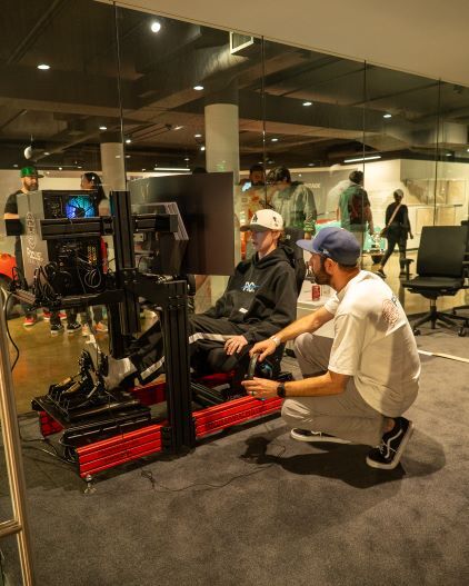 A person sits in a racing simulator with a man assisting, surrounded by other people in a gaming space.