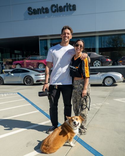 A man and woman smile together in front of a car dealership with a dog sitting at their feet.