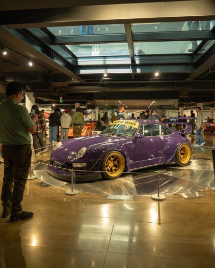 A purple Porsche 911 sits on a rotating display while people observe it in a modern museum.