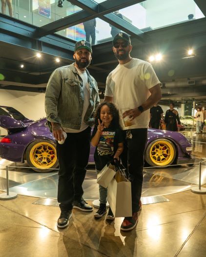 Two men and a young child pose for a photo at a car exhibit, with a purple sports car in the background.