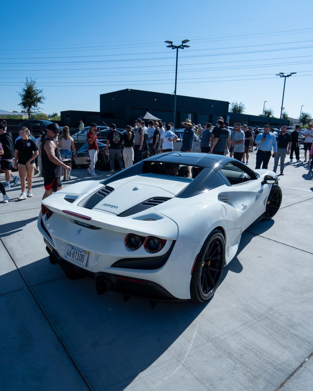 Rear view of a white Ferrari F8 Tributo parked on concrete, surrounded by a large crowd under a clear sky.