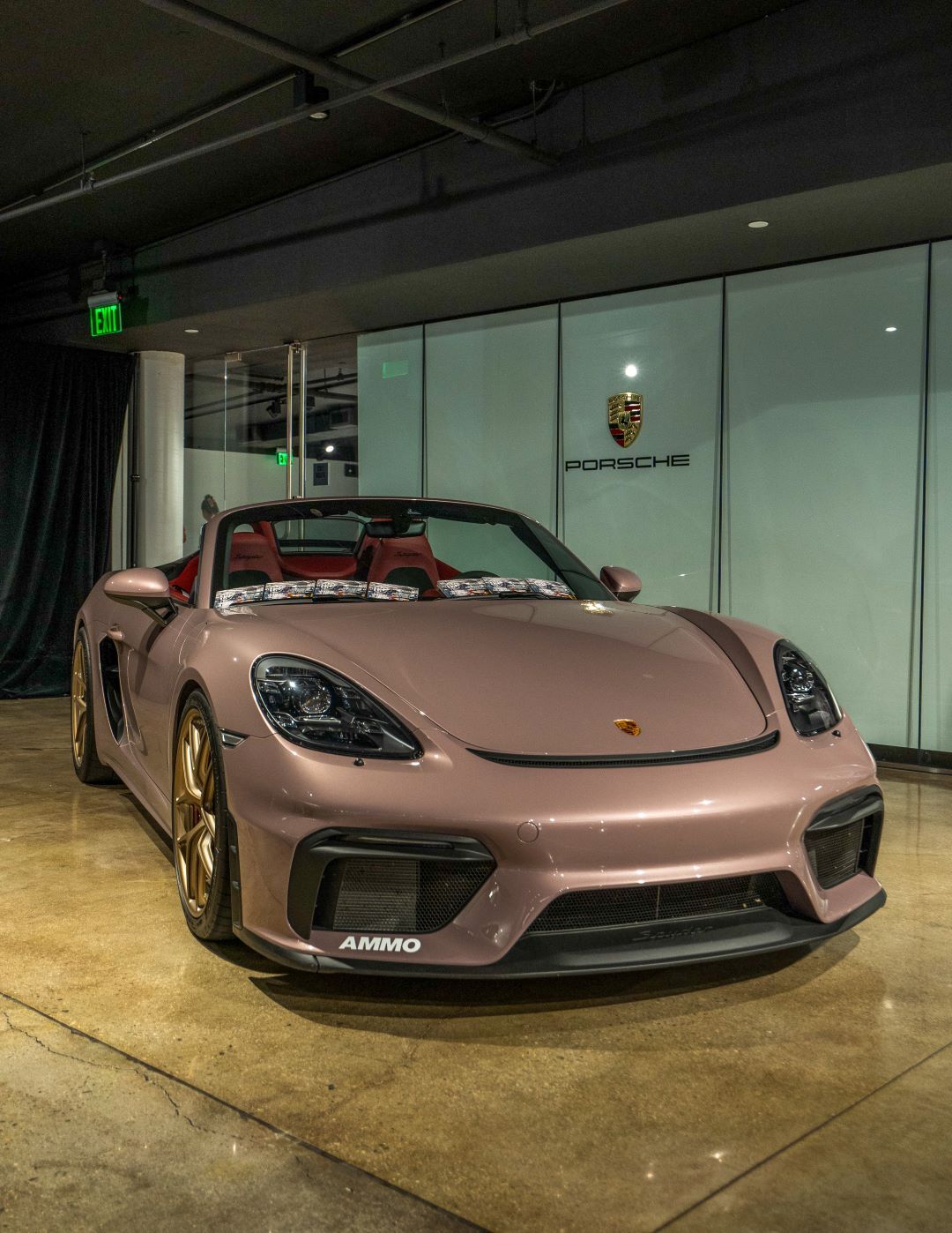 A front view of a metallic rose gold Porsche convertible with gold rims parked in a sleek garage with a Porsche logo.