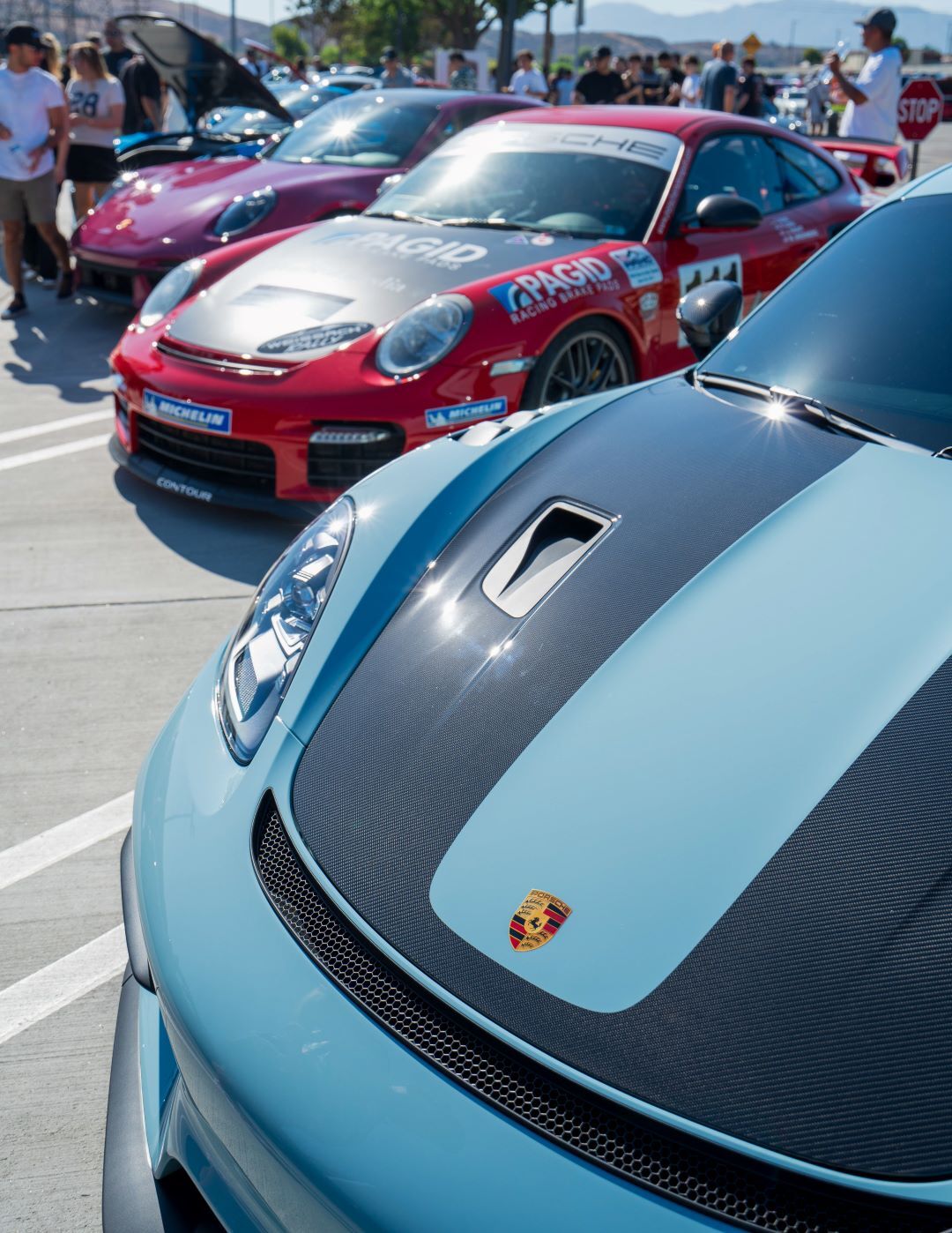 Three Porsche sports cars parked outside, including a blue car with a black stripe foreground, and red and pink cars behind it.