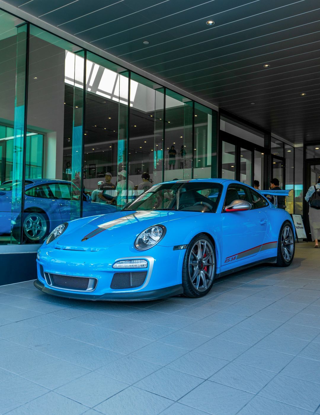 A bright blue Porsche with grey stripes and red accents is parked on a light grey tiled floor, in front of glass walls.