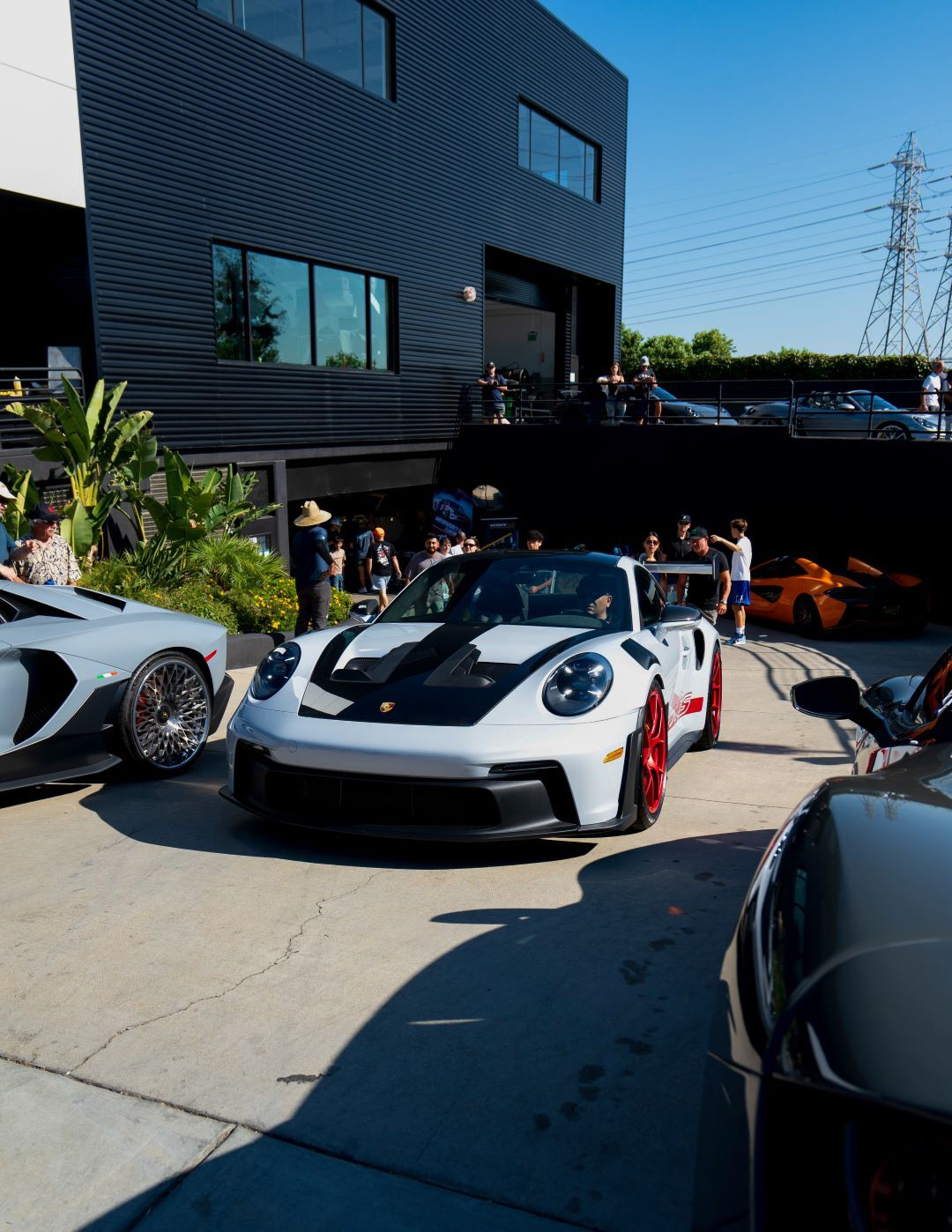 White Porsche 911 GT3 with black stripes and red wheels, next to other luxury cars outside a modern building.