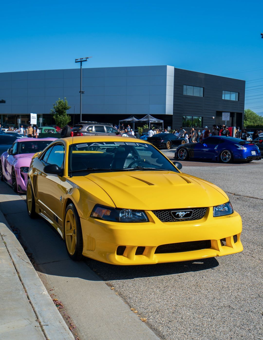 A vibrant yellow Saleen Mustang parked on a sunny day with other custom cars and a modern building in the background.