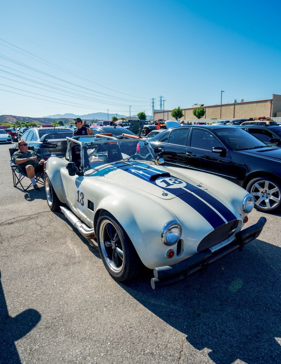 White vintage Shelby Cobra convertible with dual blue racing stripes and the number 13 parked at a car show.