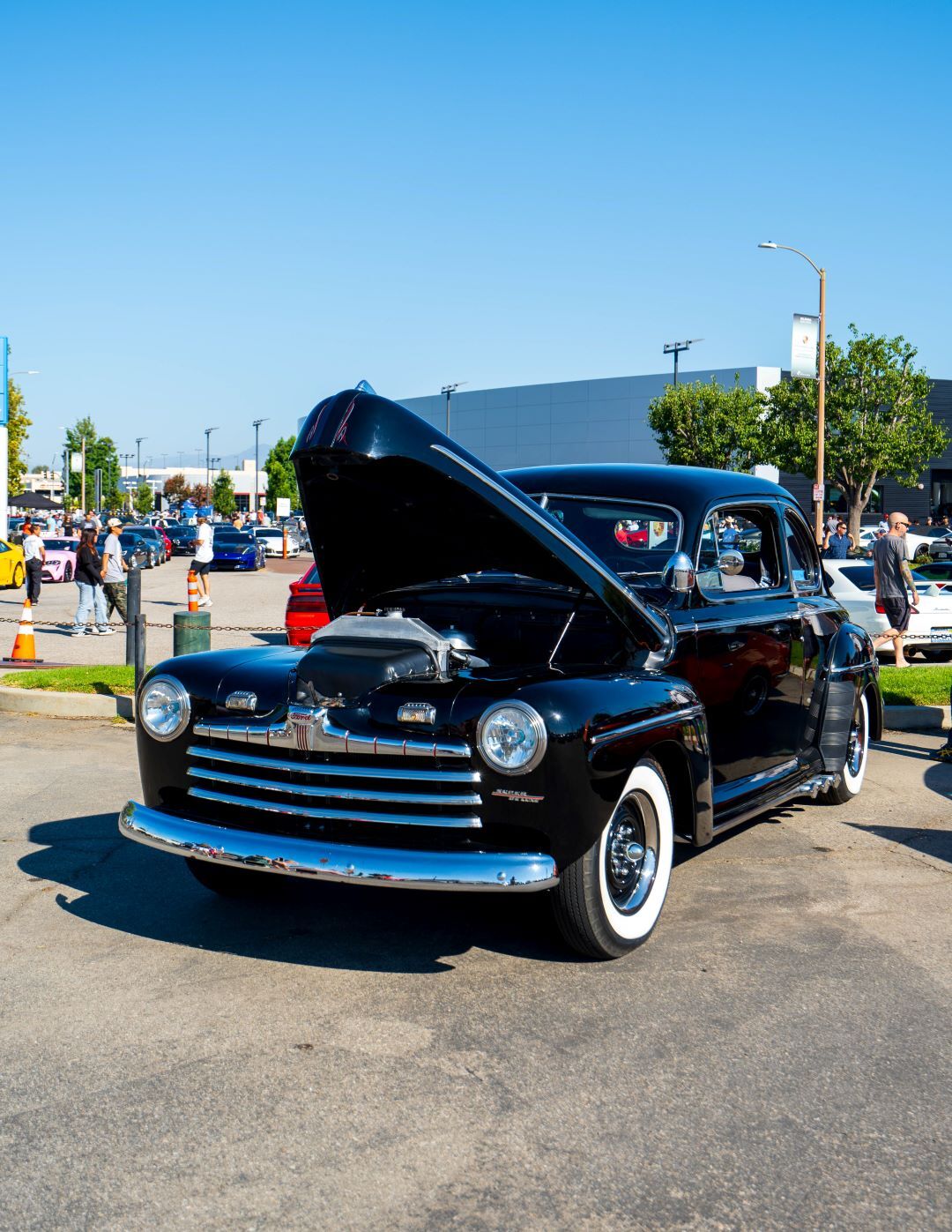 A glossy black 1940s classic car with its hood open, showcasing the engine in a sunny outdoor lot.