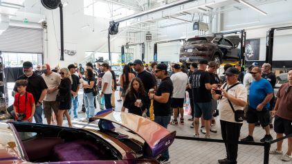 A large group of people examines a car and its parts inside a modern, well-lit garage.