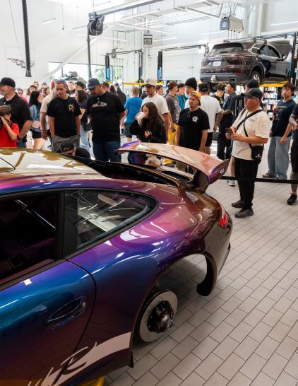 A crowd of people gathers around a colorful customized car on a lift in a garage.