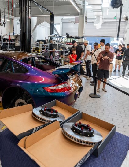 A purple Porsche sits in a garage as people observe and take photos of the car and parts.