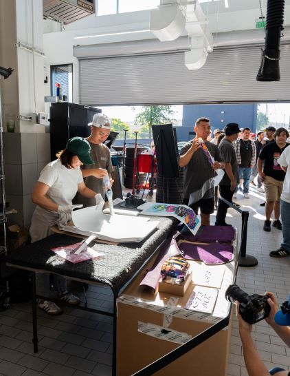 Several people look on as two technicians screen print posters at an outdoor event.