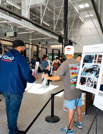 Two men in Ford Performance jackets examine a display board about Porsche restoration at a large car workshop.