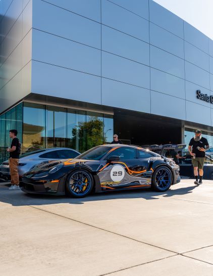 A dark blue and orange Porsche is parked in front of a modern building with two people standing nearby.