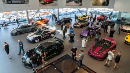 A large group of people examines a car and its parts inside a modern, well-lit garage.