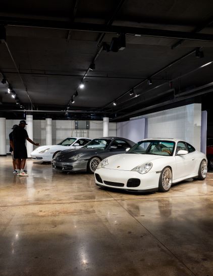 Two people stand in a dimly lit showroom surrounded by several white and gray Porsche sports cars.
