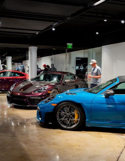 A man in a cap stands near a row of sleek, colorful Porsche sports cars displayed in a showroom.